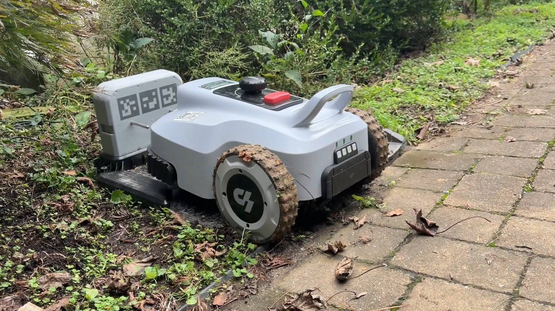 A Lopkin L2500 robot mower parked on its charging station beside a garden bed and brick walkway