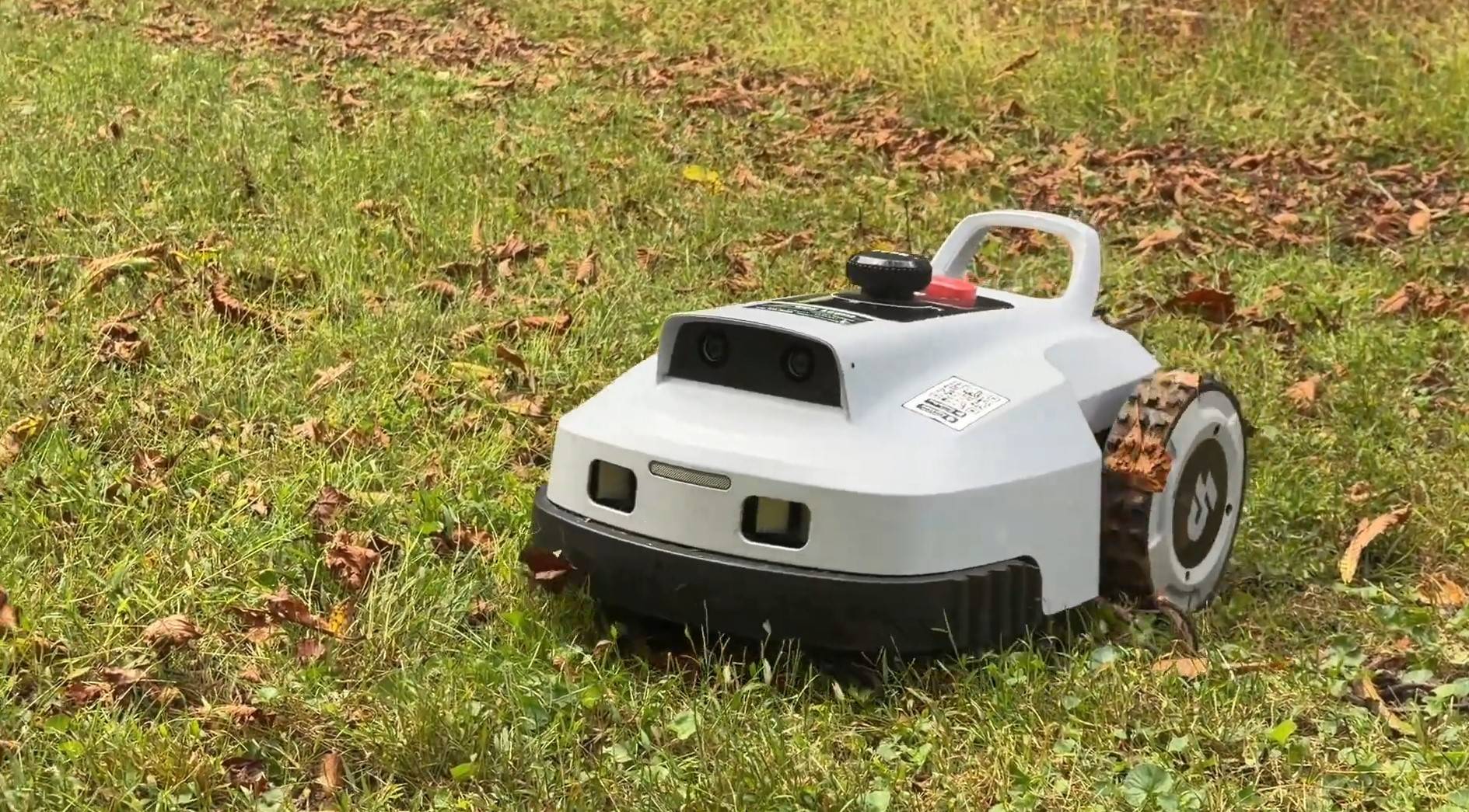 The front of a Lopkin L2500 robot mower showing its twin 3D vision cameras while mowing a lawn scattered with fall leaves