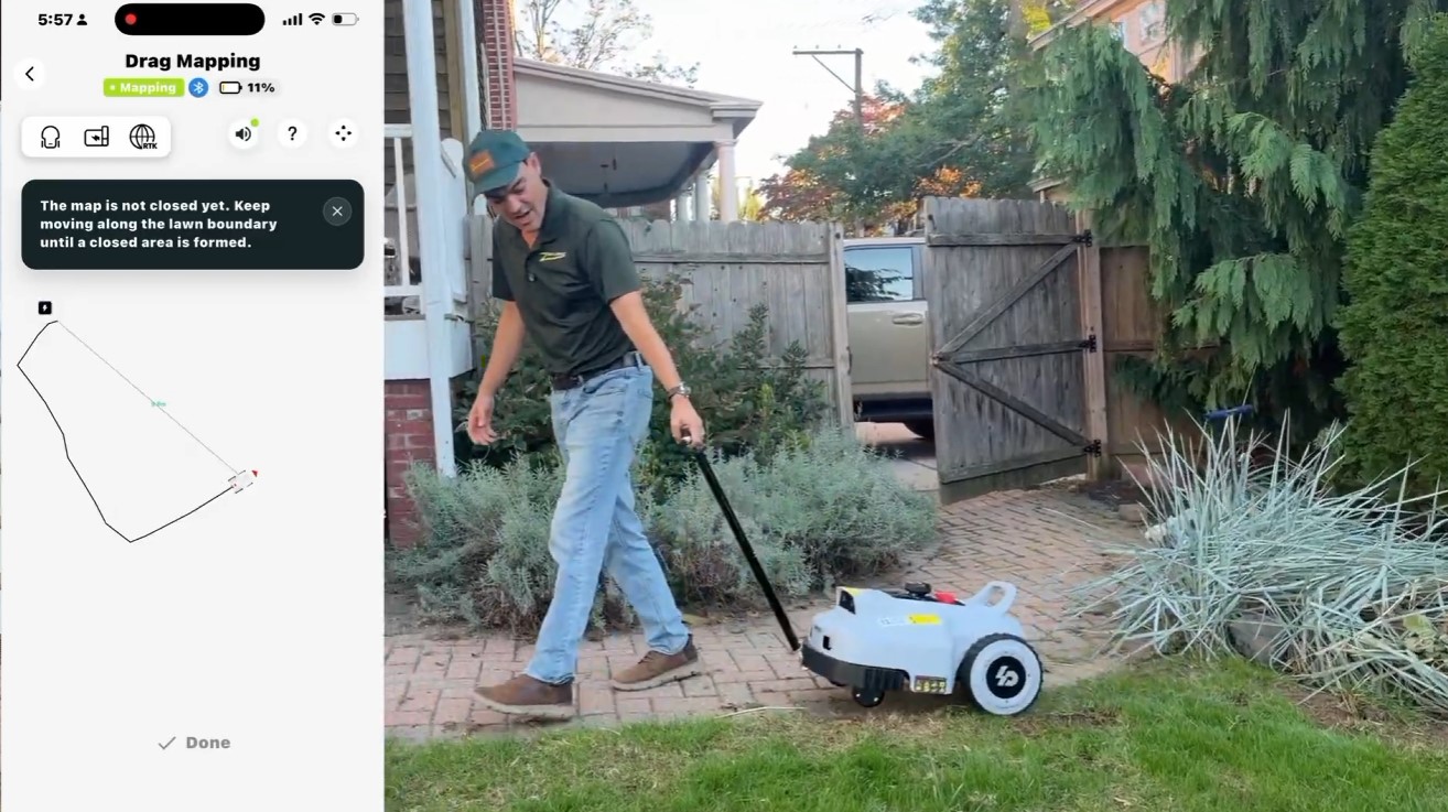 Paul pulling a Lopkin L2500 robot mower by its tow bar along a lawn edge while the app's Drag Mapping screen draws the boundary