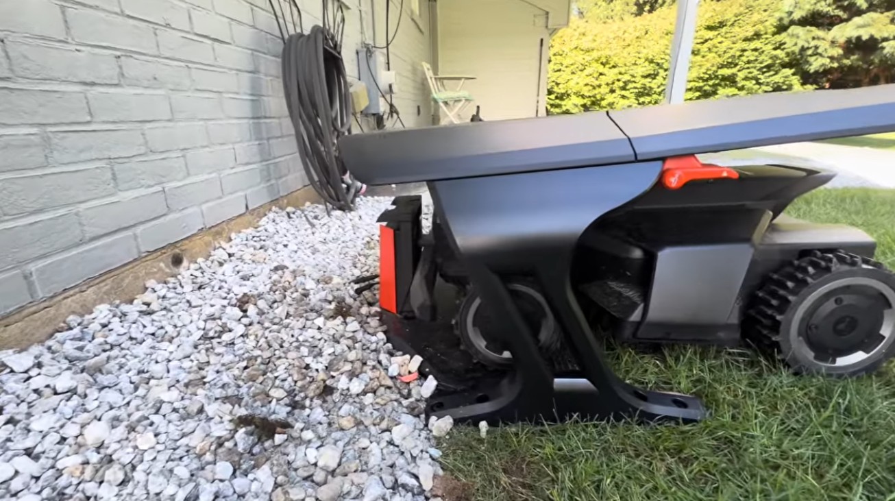 A Navimow charging station wedged against a house wall with its base overhanging a gravel bed, under the building's roofline