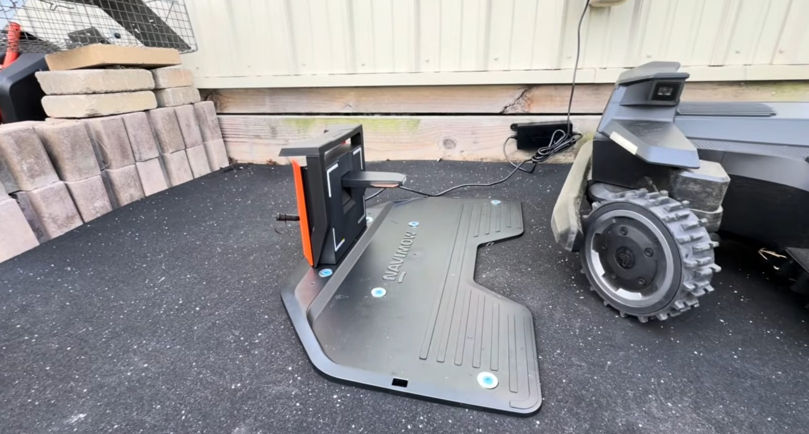 A Segway Navimow charging station sitting on a black rubber mat against a house wall, with the mower parked beside it