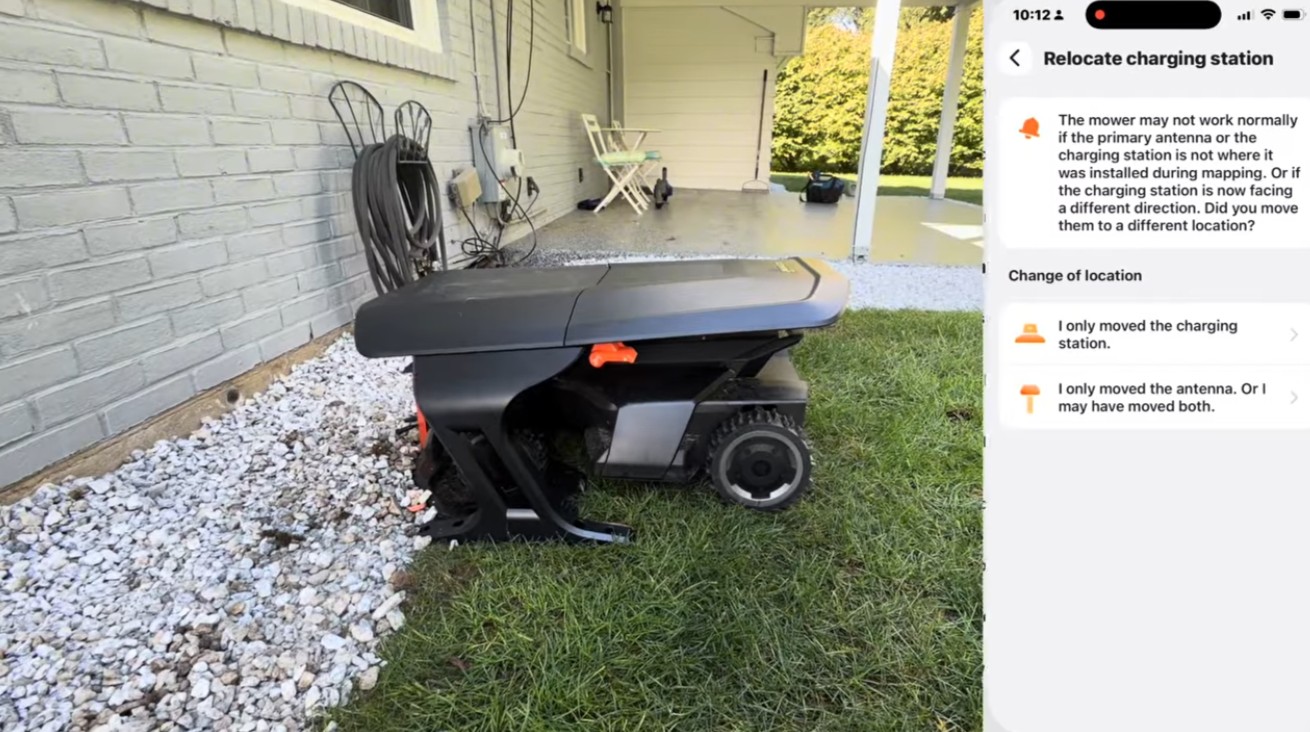 A Segway Navimow docked on its charging station beside a brick house wall, next to a phone showing the Navimow app's Relocate charging station screen