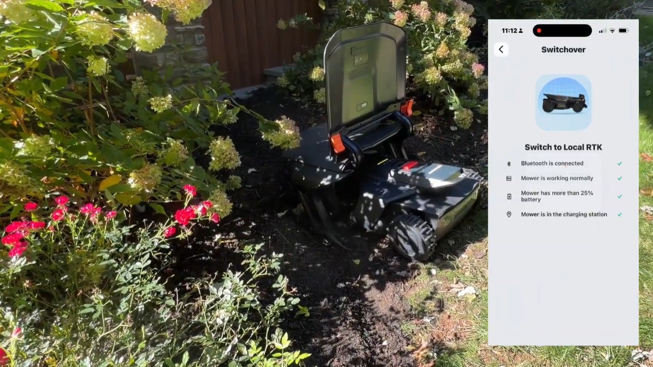 A Navimow X430 parked in its garage in a flower bed, beside a phone showing the Navimow app's Switch to Local RTK checklist