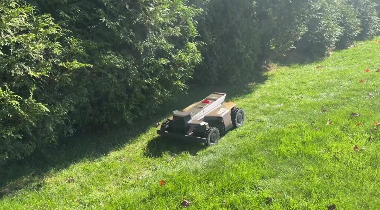 A Navimow X430 robot mower cutting a sloped lawn along a tall evergreen hedge at a Downingtown, PA home