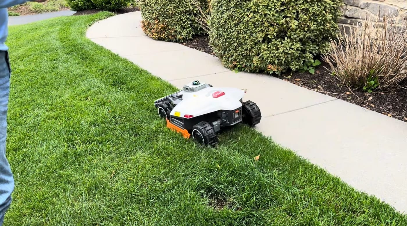 A white and orange robot mower cutting cleanly along the edge of a curved sidewalk beside a lawn