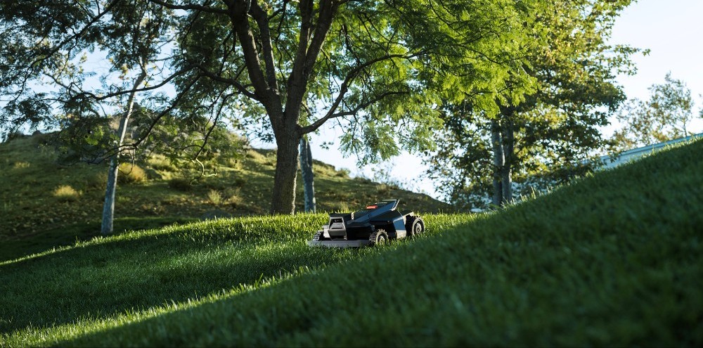 Robot mower working across a sloped, tree-lined lawn in evening light