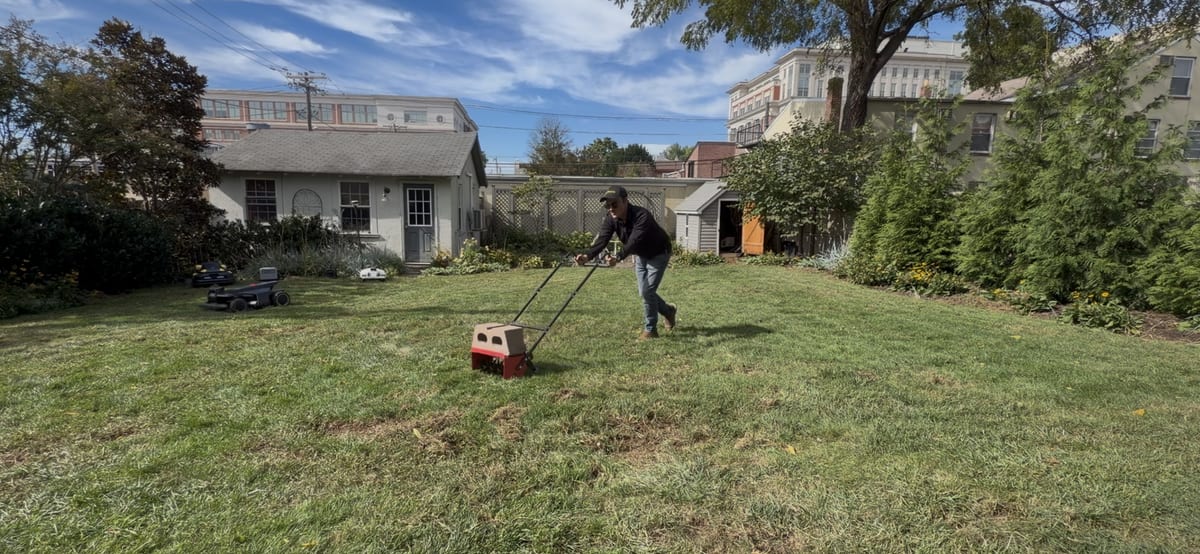Pushing a drop spreader across a freshly raked backyard lawn, a robot mower waiting behind