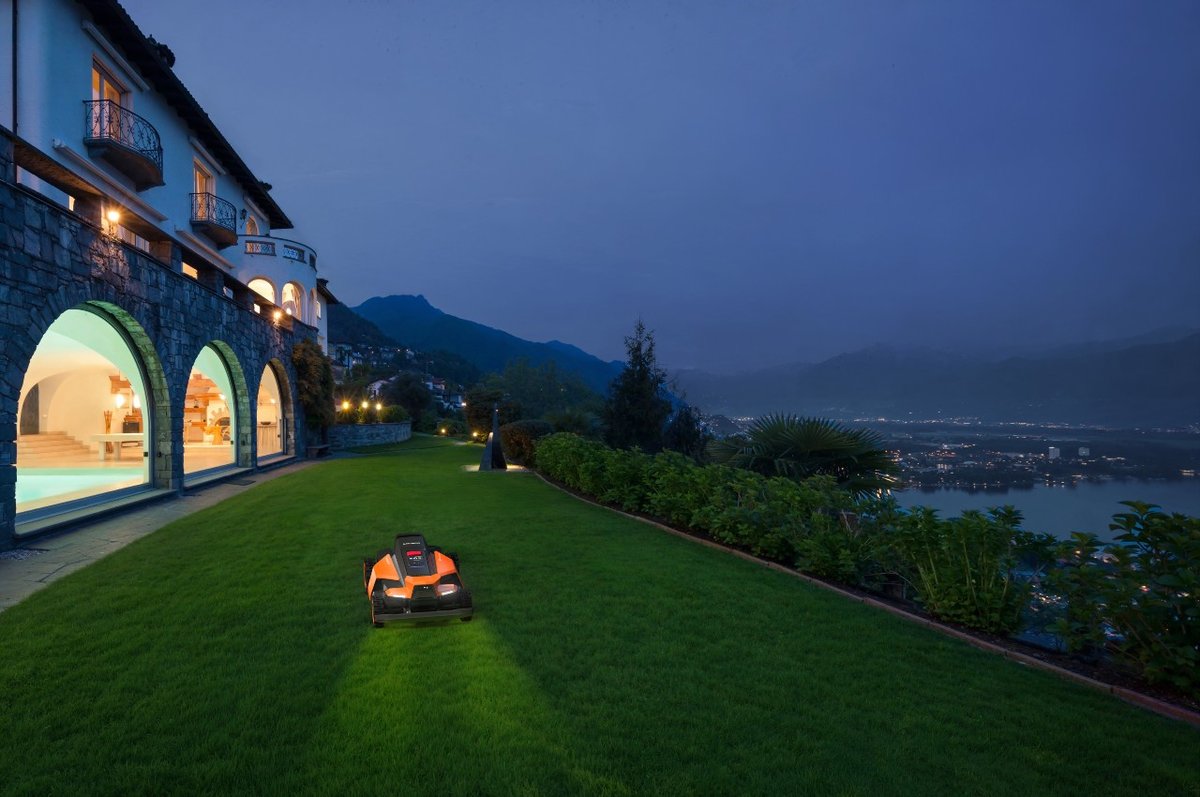 A robot mower with its headlights on cutting a hillside lawn at dusk beside a lit villa